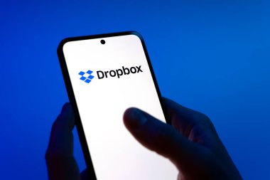 Prag, Çek Cumhuriyeti - 08 12 2025: Mavi arkaplandaki eller Dropbox logosunu gösteren bir cep telefonu tutuyor. Dropbox bulut depolama ve dosya paylaşımı için bir platformdur.