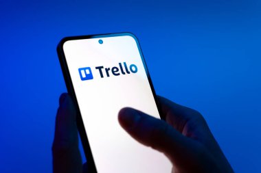 Prag, Çek Cumhuriyeti - 08 12 2025: Mavi arkaplandaki eller Trello logosunu gösteren bir cep telefonu tutuyor. Trello, tahta ve görevleri kullanarak proje yönetimi için bir platformdur.