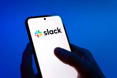 Prag, Çek Cumhuriyeti - 08 12 2025: Mavi arkaplandaki eller Slack logosunu gösteren bir cep telefonu tutuyor. Slack takım mesajlaşması ve iş yeri iletişimi için bir platformdur.