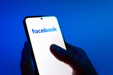 Prag, Çek Cumhuriyeti - 08 12 2025: Eller mavi arkaplanda Facebook logosunu gösteren bir cep telefonu tutuyor. Facebook, içeriği bağlamak ve paylaşmak için bir sosyal medya platformudur.