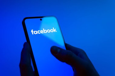 Prag, Çek Cumhuriyeti - 08 12 2025: Eller mavi arkaplanda Facebook logosunu gösteren bir cep telefonu tutuyor. Facebook, içeriği bağlamak ve paylaşmak için bir sosyal medya platformudur.