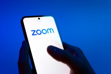 Prag, Çek Cumhuriyeti - 08 12 2025: Mavi arkaplandaki eller Zoom logosunu gösteren bir cep telefonu tutuyor. Zoom video toplantıları ve çevrimiçi işbirliği için bir platform.