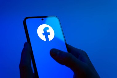Prag, Çek Cumhuriyeti - 08 12 2025: Eller mavi arkaplanda Facebook logosunu gösteren bir cep telefonu tutuyor. Facebook, içeriği bağlamak ve paylaşmak için bir sosyal medya platformudur.