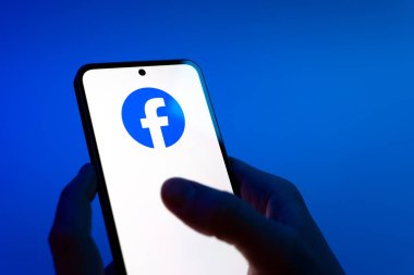 Prag, Çek Cumhuriyeti - 08 12 2025: Eller mavi arkaplanda Facebook logosunu gösteren bir cep telefonu tutuyor. Facebook, içeriği bağlamak ve paylaşmak için bir sosyal medya platformudur.