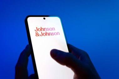 Prag, Çek Cumhuriyeti - 08 12 2025: Eller mavi arkaplanda Johnson ve Johnson logosunu gösteren bir cep telefonu tutuyor. Johnson ve Johnson, sağlık ve tüketim ürünleri üreten bir şirket..