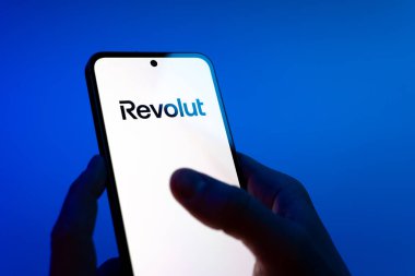 Prague, Czechia - 08 12 2025: Hands on a blue background are holding a mobile phone displaying the Revolut logo. Revolut is a platform for digital banking and financial services.