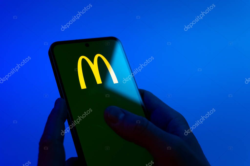 Prague, Czechia - 08 12 2025: Hands on a blue background are holding a mobile phone displaying the McDonalds logo. McDonalds is a fast-food chain serving burgers, fries, and more.