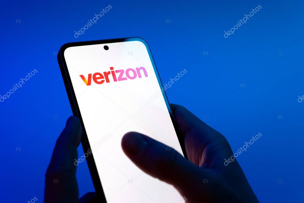 Prague, Czechia - 08 12 2025: Hands on a blue background are holding a mobile phone displaying the Verizon logo. Verizon is a telecom company providing wireless and broadband services.