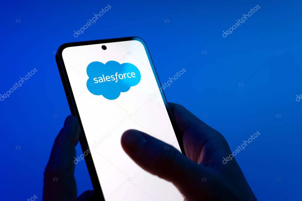 Prague, Czechia - 08 12 2025: Hands on a blue background are holding a mobile phone displaying the Salesforce logo. Salesforce is a platform for customer relationship management and cloud services.