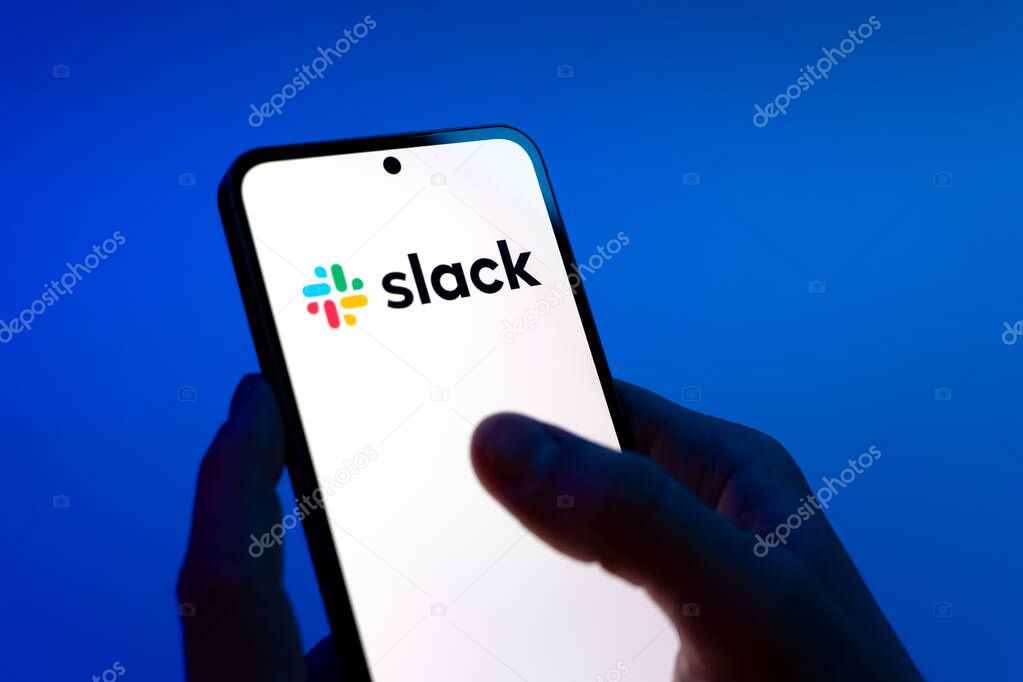 Prague, Czechia - 08 12 2025: Hands on a blue background are holding a mobile phone displaying the Slack logo. Slack is a platform for team messaging and workplace communication.