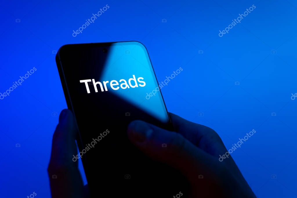 Prague, Czechia - 08 12 2025: Hands on a blue background are holding a mobile phone displaying the Threads logo. Threads is a social media platform for sharing short posts and updates.