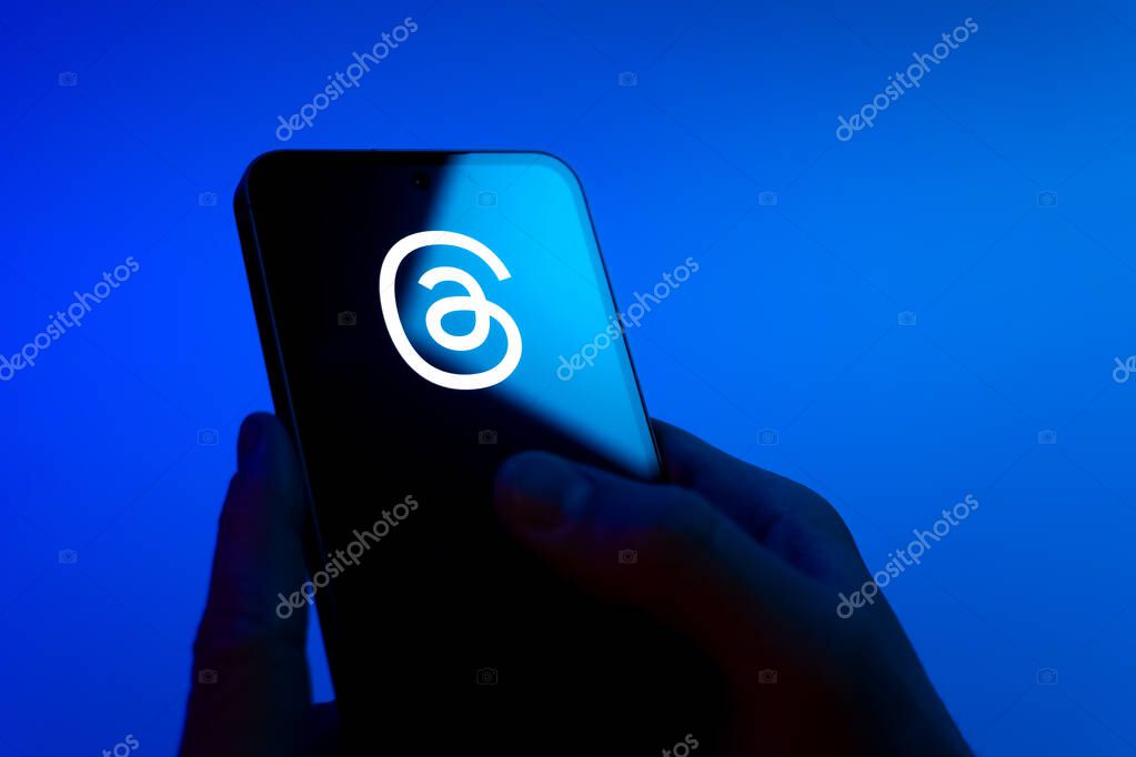Prague, Czechia - 08 12 2025: Hands on a blue background are holding a mobile phone displaying the Threads logo. Threads is a social media platform for sharing short posts and updates.