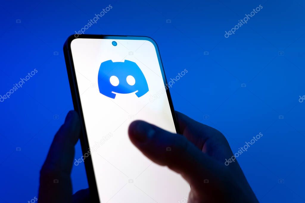Prague, Czechia - 08 12 2025: Hands on a blue background are holding a mobile phone displaying the Discord logo. Discord is a platform for voice, video, and text communication.