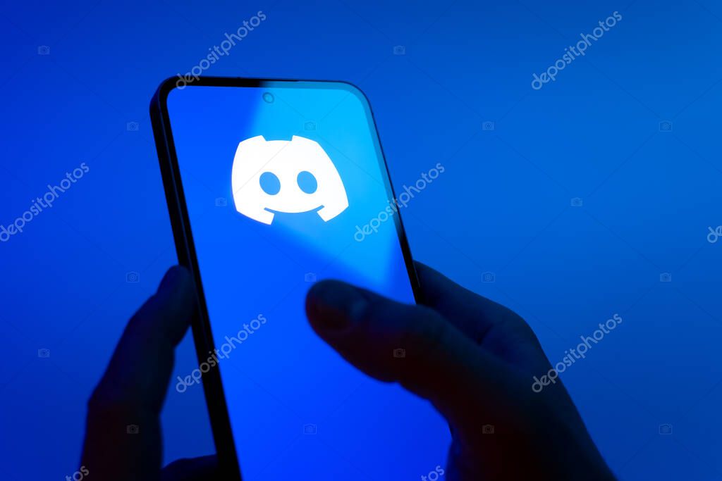 Prague, Czechia - 08 12 2025: Hands on a blue background are holding a mobile phone displaying the Discord logo. Discord is a platform for voice, video, and text communication.