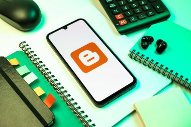 Prag, Çek Cumhuriyeti - 08 18 2025: Blog logosunu gösteren bir cep telefonuyla ofis malzemeleri düzenlemesi. Blogger blog oluşturma ve yayınlama için bir platformdur.