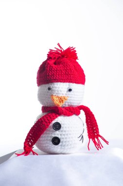 Kırmızı şapkalı ve eşarplı küçük beyaz bir kardan adam. Kardan adam beyaz bir yüzeyde oturuyor, muhtemelen bir masa ya da tezgah.