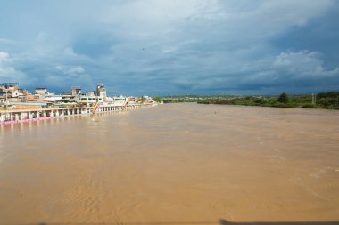 Bir nehir kahverengi ve çamurludur, arka planda birkaç ev görülebilir. Gökyüzü bulutlu, ortama kasvetli ve kasvetli bir hava veriyor.