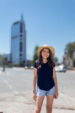 Hasır şapkalı ve mavi gömlekli bir kadın yüksek bir binanın önünde duruyor. Gülümsüyor ve eğleniyor.