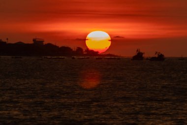Güneş okyanusun üzerinde batıyor, suyun üzerine sıcak bir parıltı saçıyor. Gökyüzü bulutlarla dolu, sakin ve huzurlu bir atmosfer yaratıyor. Sudaki tekneler küçük ve dağınık.