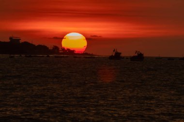Güneş okyanusun üzerinde batıyor, suyun üzerine sıcak bir parıltı saçıyor. Gökyüzü bulutlarla dolu, sakin ve huzurlu bir atmosfer yaratıyor. Sudaki tekneler küçük ve dağınık.
