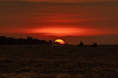 Güneş okyanusun üzerinde batıyor, suyun üzerine sıcak bir parıltı saçıyor. Gökyüzü bulutlarla dolu, sakin ve huzurlu bir atmosfer yaratıyor. Sudaki tekneler küçük ve dağınık.