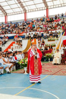 Tumbes, Tumbes, Peru 21-10-2023, Kuvvetlendirme Ayini: Kırmızı cüppeli bir adam kalabalığın önünde duruyor. Elinde bir mikrofon var ve konuşma yapıyor. Seyirciler arasında pek çok kişinin olduğu atmosfer canlı ve enerjik.