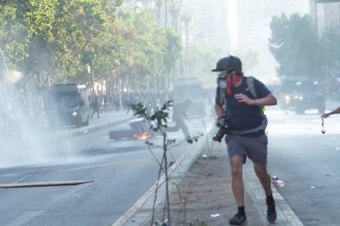 Santiago de Chile - Şili, 25-11-2019: Mavi tişörtlü ve gri şortlu bir adam gaz maskesi takmış bir şekilde caddede koşuyor. Cadde duman ve molozlarla dolu. Yolun kenarında park etmiş birkaç araba ve kamyon var.