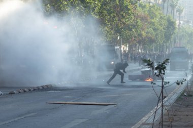 Santiago de Chile, Şili, 25-11-2019: Bir adam arabanın camını kırmaya çalışıyor. Sahne kaotik ve şiddet dolu. Arka planda bir sürü insan var, bazıları sırt çantası taşıyor. Ateşten çıkan duman çok yoğun.