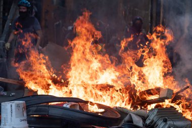 Lastik ve odun yığınında bir ateş yanıyor. Siyah gömlekli bir adam ateşin arkasında duruyor.