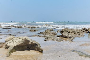 Önünde büyük bir kaya olan kayalık bir sahil. Gökyüzü açık ve su sakin.