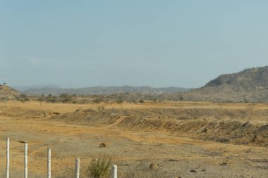 Çorak bir çöl manzarası, birkaç ağaç ve birkaç insan var. Gökyüzü açık ve güneş parlıyor.