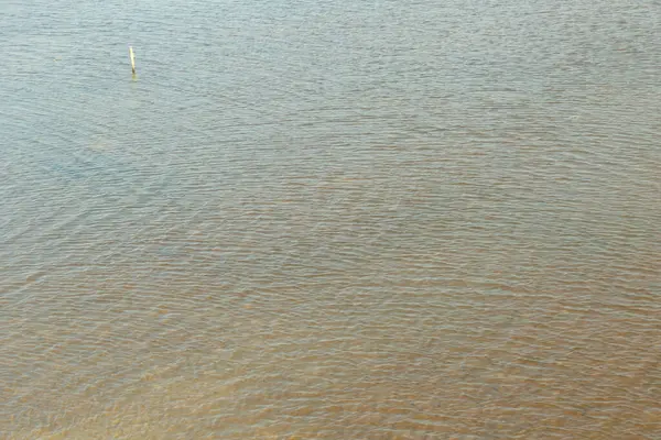 Su sakin ve hareketsiz, yüzeyde dalgalanmalar oluşuyor. Gökyüzü bulutlu ve su açık kahverengi bir renk gibi görünüyor.