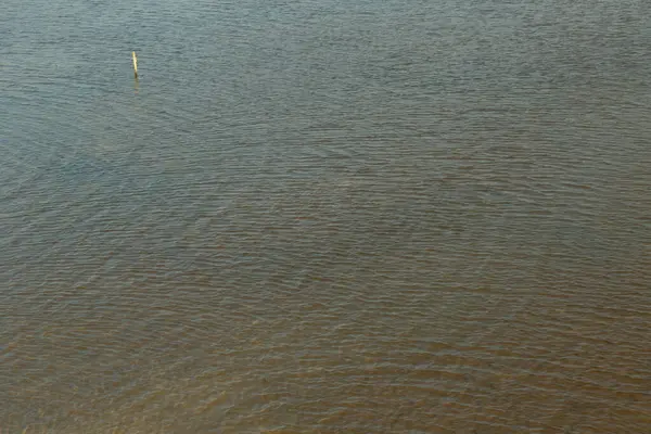 Su sakin ve hareketsiz, yüzeyde dalgalanmalar oluşuyor. Gökyüzü bulutlu ve su bulanık görünüyor.