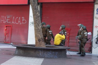 Santiago de Chile - Şili, 20-10-2019: Bir adam bir ağacın önünde diz çöküyor ve arkasında bir grup polis memuru duruyor.