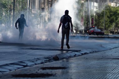 Santiago de Chile - Şili, 20-10-2019: İki adam dumanlı ve arkalarında bir arabayla caddede koşuyor. Sahne kaotik ve yoğun.