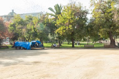 Kamyonunda mavi muşamba olan bir park ve banklarda oturan birkaç kişi. Park boş ve ağaçlar yeşil.