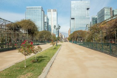 Santiago de Chile, Şili, 27-06-2021: Ortasında park olan bir şehir sokağı. Park binalarla çevrili ve kaldırımı var. Parkta kırmızı çiçekli olan da dahil olmak üzere birkaç ağaç var. Sokak bomboş..
