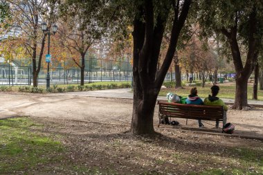 Santiago de Chile - Şili, 27-06-2021: Parktaki bir bankta oturan iki kişi. Park ağaçlarla çevrili ve arka planda bir basketbol sahası var. Manzara huzurlu ve rahatlatıcı..