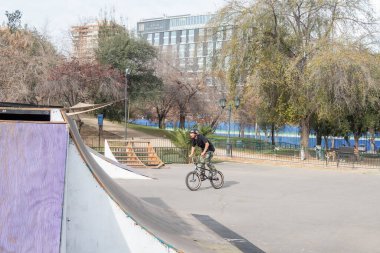 Santiago de Chile, Şili, 27-06-2021: Bir adam parkta bisiklet sürüyor. Park binalarla çevrili ve bir kaykay parkı var..