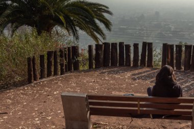 Parktaki bankta oturan bir kadın. Bankın etrafı tahta direklerle çevrili ve kadın şehre bakıyor. Manzaranın tadını çıkaran kadınla huzur dolu ve huzurlu bir sahne.