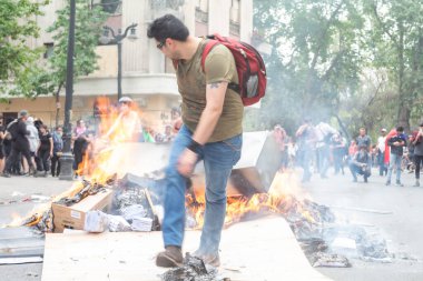 Santiago de Chile - Şili, 25-10-2019: Yeşil gömlekli ve kot pantolonlu bir adam bir yığın kağıt ve karton üzerinde yürüyor. Sahne kaotik ve şiddetli, insanlar koşuyor ve bağırıyor. Adam kaostan kaçmaya çalışıyor.