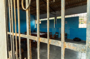 Demir pencereli ve metal kapılı bir hapishane hücresi. Oda boş ve mavi bir duvarı var.