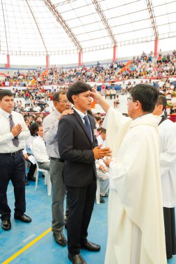 Bir adam kalabalığın önünde bir rahip tarafından kutsandı. Rahip beyaz bir bornoz giyiyor ve kutsanmış adam da takım elbise giyiyor.