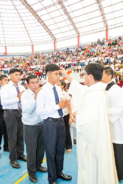 Bir adam kalabalığın önünde genç bir adamı kutsuyor. Adam beyaz bir bornoz giyiyor ve genç adam beyaz bir gömlek ve kravat takıyor. Kalabalık töreni izliyor.