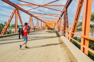 Tumbes, Tumbes, Peru 01-03-2025: Kırmızı gömlekli bir adam arkasında bir kamyonla köprüden geçiyor. Köprü turuncu ve gökyüzü mavi.