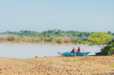 Nehirdeki küçük bir teknede iki kişi oturuyor. Gökyüzü açık ve güneş parlıyor.