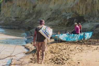 Sırtında bir çantayla sahilde yürüyen bir adam var. Arkasında bir tekne var.