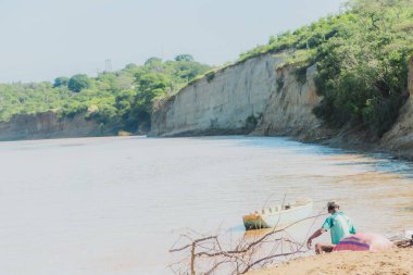 Nehirdeki bir teknede bir adam oturuyor. Su kahverengi ve bulanık. Adam balık tutuyor.