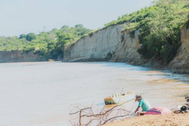 Sudaki bir teknede bir adam oturuyor. Su kahverengi ve bulanık. Adam yerde bir şeye bakıyor.