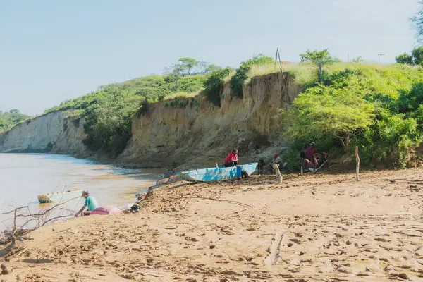 Bir grup insan sahile yakın oturuyorlar. Kumsal kayalık ve su bulanık.